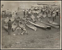 Two rows of canoes sit on a grassy beach behind a crowd of spectators and, in front of them in the background, open water.